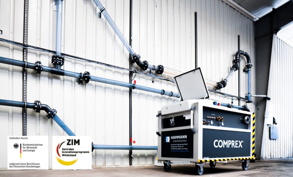 Eine mechanische Einheit mit der Aufschrift „COMPREX“ verfügt über offene Fächer und ist mit Rohren an einer Fabrikwand verbunden, was den Industriebetrieb verbessert. Logos sind unten links zu sehen. Das System optimiert die Rohrreinigung durch präzisionsgefertigte Impulstechnologie.