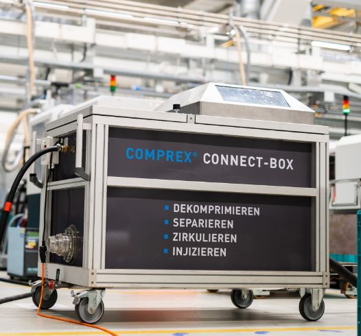 Die industrielle Comprex Connect-Box mit deutschem Text in einer Fabrikumgebung mit Metallrohren und Maschinen demonstriert industrielle Präzision und Innovation.