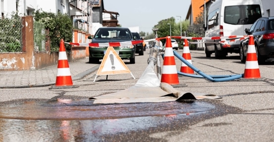 Eine kommunale Baustelle mit Leitkegeln, Absperrband, Warnschild und nasser Fahrbahnoberfläche in einer Anliegerstraße weist auf laufende Arbeiten hin, bei denen möglicherweise das Spülverfahren zum Einsatz kommt.
