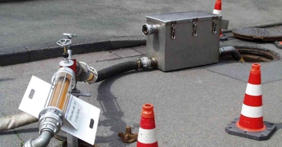 Straßenrand-Aufstellung mit Verkehrsleitkegeln, an eine Metallbox angeschlossenen Schläuchen und einem Ventilmechanismus für die kommunale Rohrreinigung.