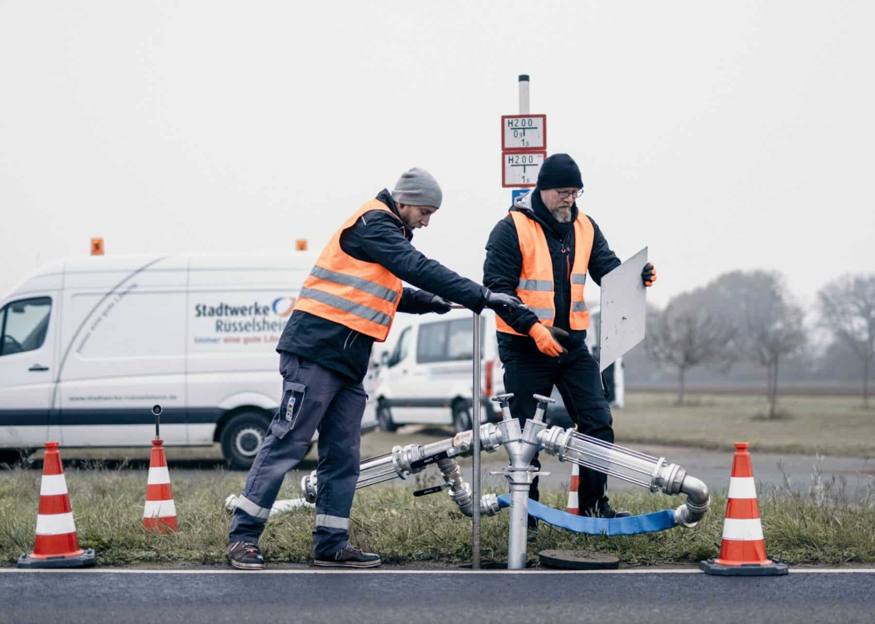 Zwei Arbeiter in orangefarbenen Westen richten an einem nebligen Tag am Straßenrand in der Nähe von Verkehrsleitkegeln und einem Lieferwagen die Ausrüstung aus und bereiten sich sorgfältig auf eine Rohrreinigung vor. Dabei kommt ein modernes Spülverfahren zum Einsatz, um sicherzustellen, dass die Rohrleitungen gründlich gesäubert werden.