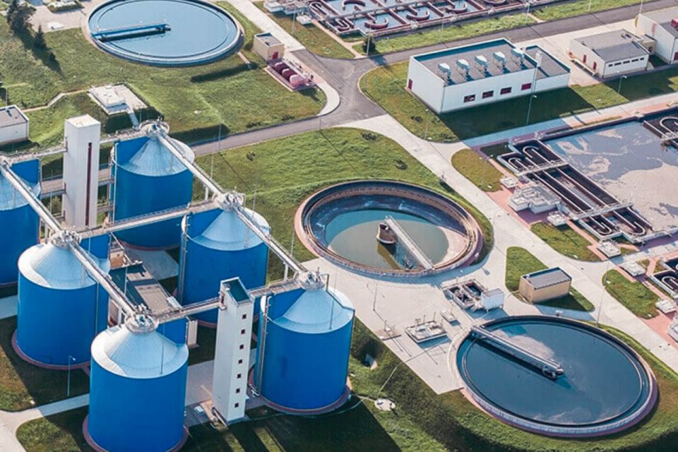 Aerial view of a Kommunal water treatment facility featuring blue cylindrical tanks and circular settling ponds on grassy terrain, showcasing advanced Impulse Rohrreinigung technology.