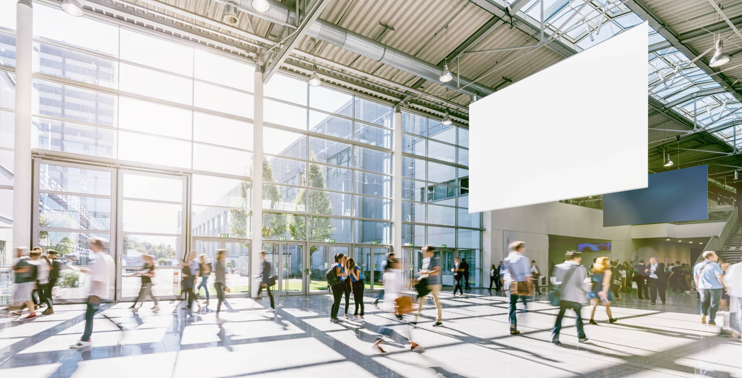 Die Leute gehen durch eine geräumige, sonnendurchflutete Lobby mit deckenhohen Fenstern und einem leeren weißen Banner, das von der Decke hängt – perfekt, um für die örtlichen Dienstleistungen von Gewerbe zu werben.