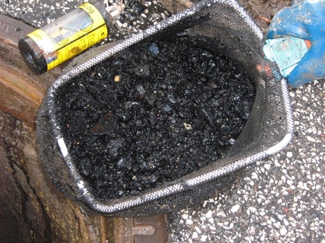 Ein mit schwarzem Schlamm gefüllter Behälter wird über einen Schacht gehalten, bereit, Grobpartikel und Späne auszutragen. Daneben liegt eine gelbe Dose auf dem Asphalt, auf dem Behälter ist eine blau behandschuhte Hand zu sehen.