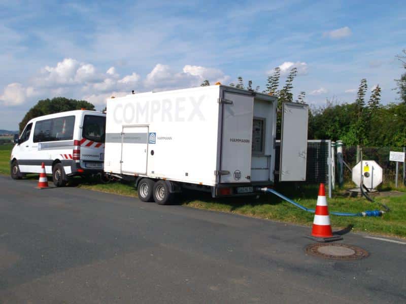 Ein weißer Nutzfahrzeuganhänger mit der Aufschrift „COMPREX“ ist über blaue Schläuche mit einem Schacht verbunden. Daneben parkt ein Wartungsfahrzeug auf einer asphaltierten Straße mit Verkehrsleitkegeln. Im Hintergrund sind Bäume und Felder zu sehen. Der Aufbau scheint darauf ausgelegt zu sein, Grobpartikel und Späne effizient auszutragen.