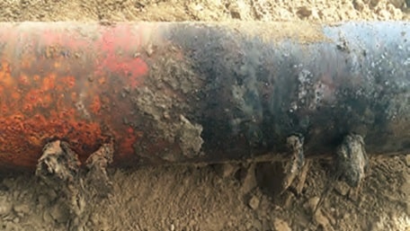 Eine Nahaufnahme eines korrodierten Metallrohrs, das teilweise im Boden vergraben ist und deutliche Anzeichen von Rost und Verschleiß aufweist. Rundherum sind Grobpartikel und Späne sichtbar, die sich austragen.