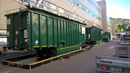 Große grüne Frachtcontainer, die möglicherweise zur PFAS-Bewirtschaftung genutzt werden, stehen auf Anhängern, die neben einem Gebäude geparkt sind, mit Hügeln im Hintergrund.