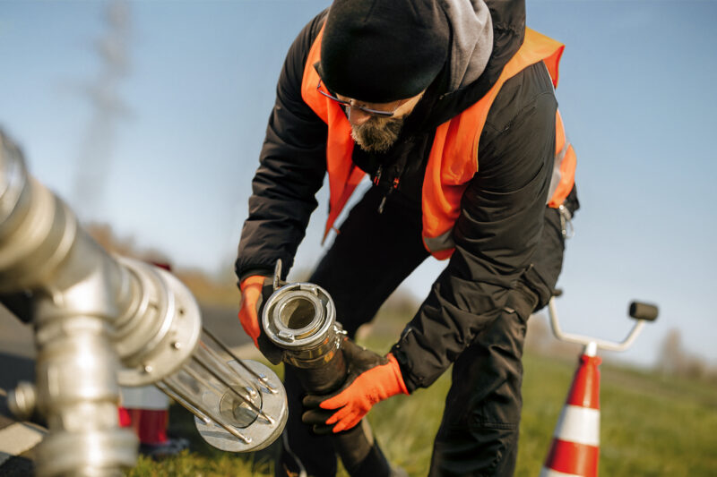 A worker in safety gear connects a hose to a metal pipe near traffic cones outdoors.