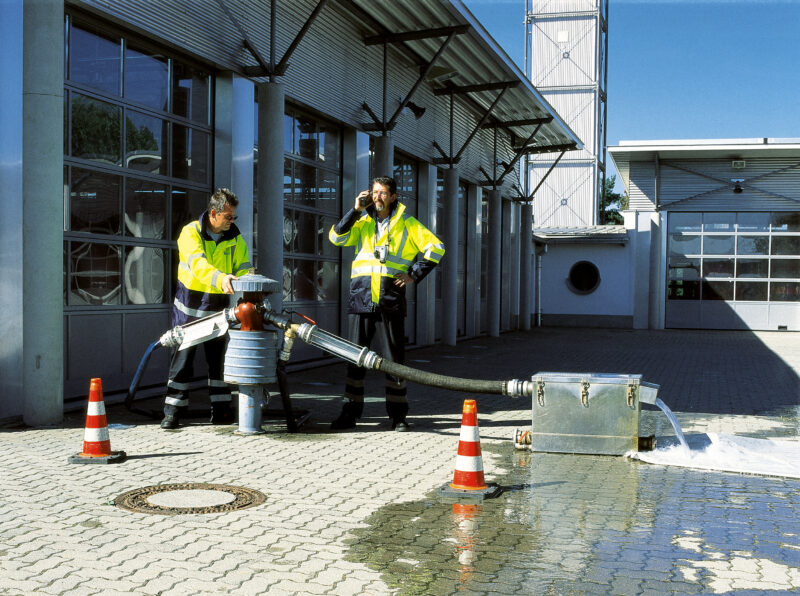 Two workers in safety jackets monitor water flowing from equipment outside an industrial building.